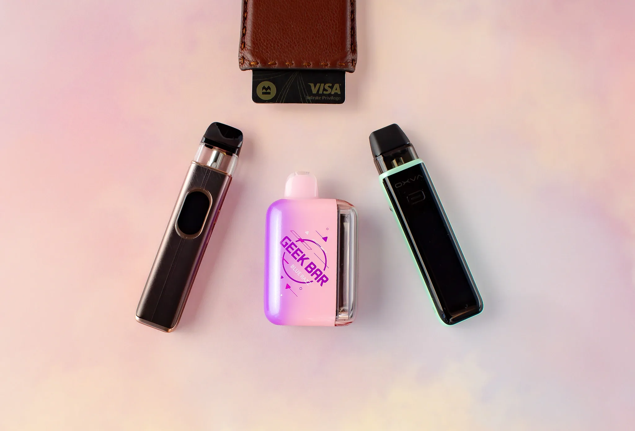 Three disposable pod vapes in various colors arranged around a brown wallet with a credit card on a soft background.